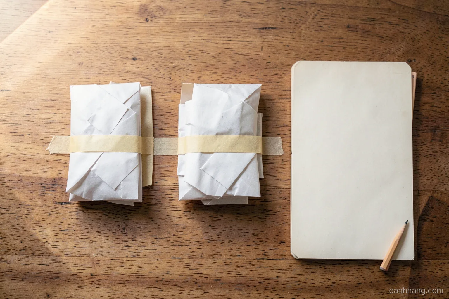 Top-down view of a wooden table with three neat piles of small unmarked parcels separated by masking tape lines, a plain notebook and a pencil, soft morning light from the left