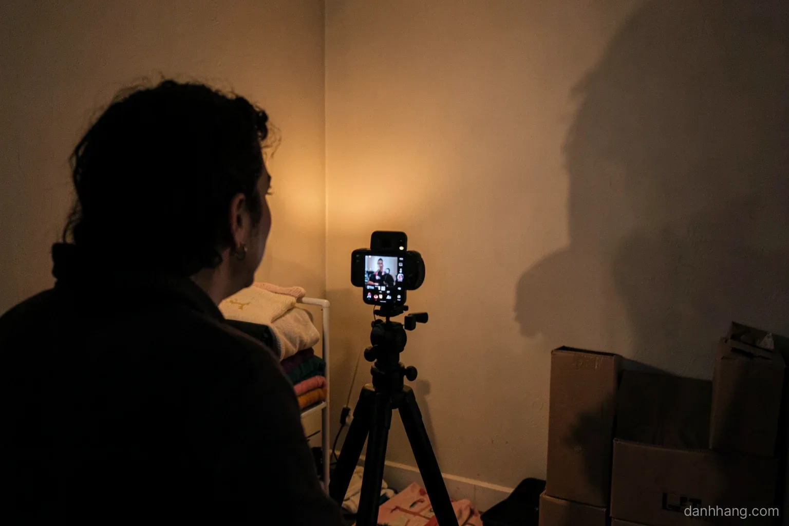 A small home livestream corner at night seen from behind the seller, a phone on a tripod facing a plain wall with folded sweaters and boxes on a rack, warm lamp light